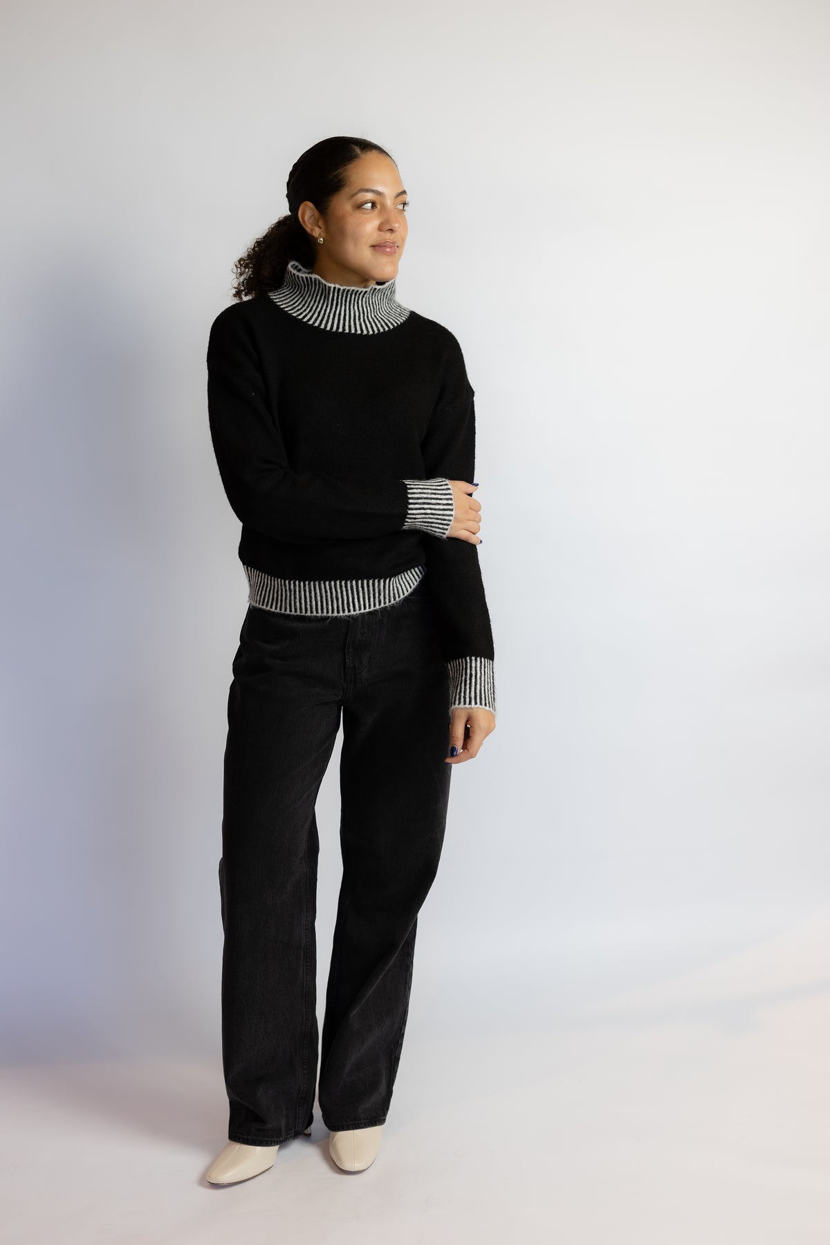 A woman modeling a black knit sweater with a black and white striped turtleneck collar, cuffs, and hem, paired with black pants and off-white pointed shoes, against a plain light gray studio background.