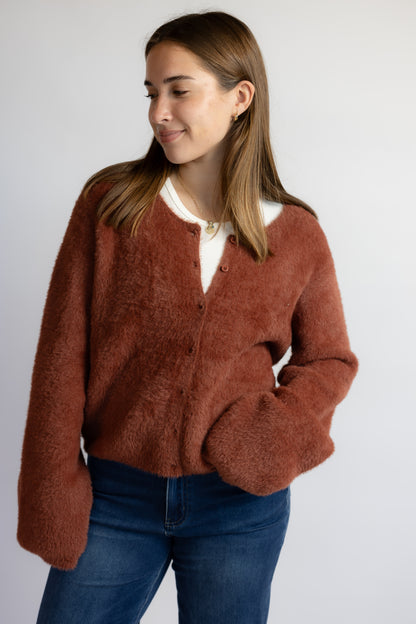 A woman wearing a fuzzy, textured rust-brown cardigan over a white collared shirt and blue jeans, photographed against a plain light gray studio background.