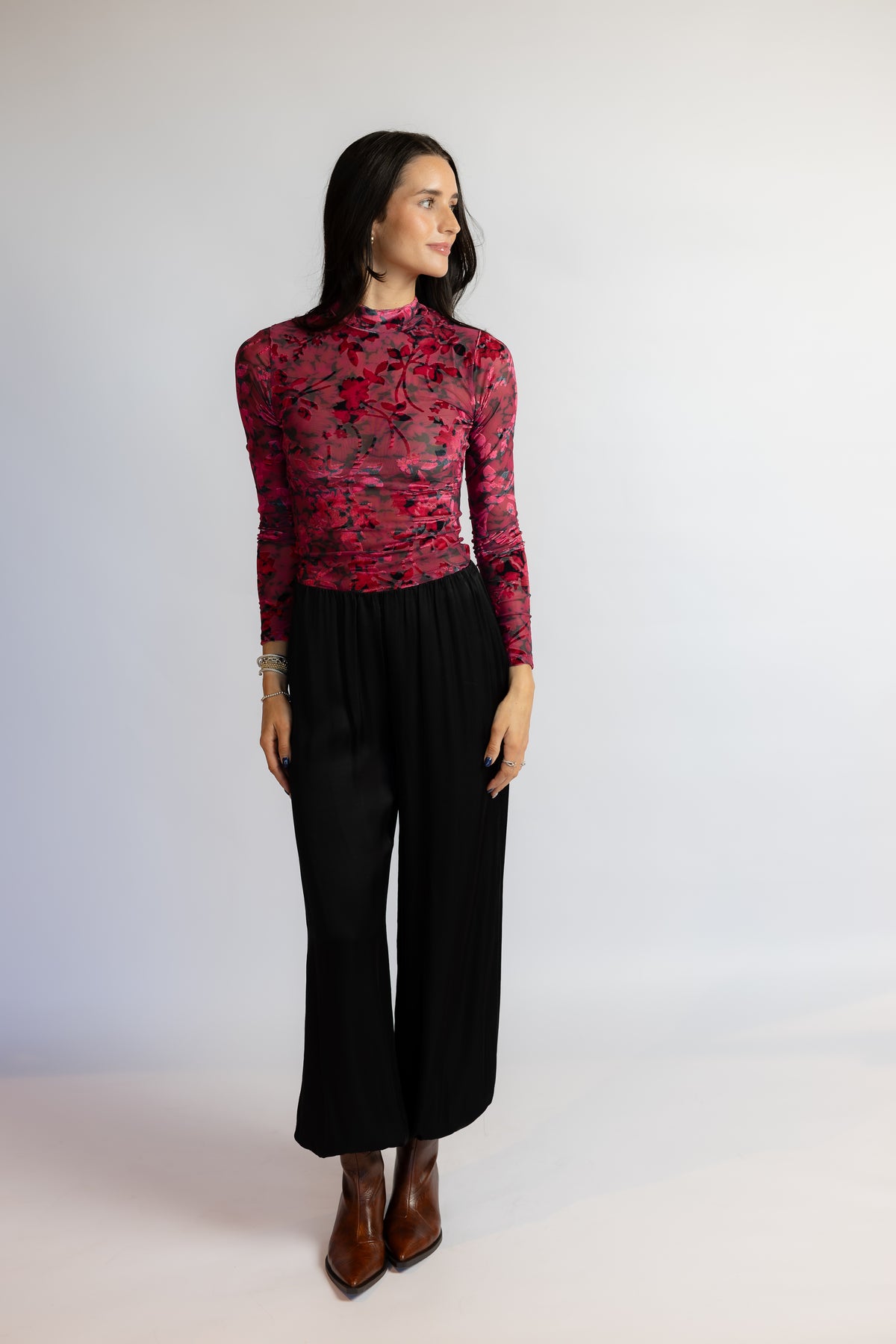 A woman modeling a long-sleeve mock neck top with a red and black floral lace overlay, paired with black wide-leg pants and brown ankle boots, against a plain light gray studio background.