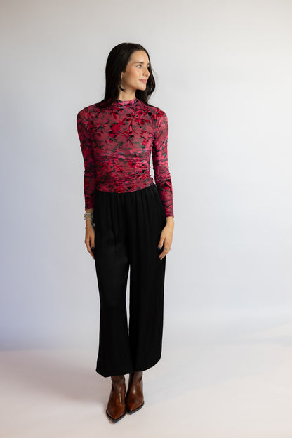 A woman modeling a long-sleeve mock neck top with a red and black floral lace overlay, paired with black wide-leg pants and brown ankle boots, against a plain light gray studio background.