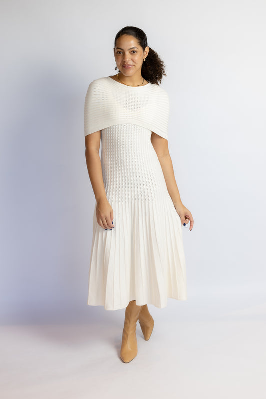 A woman modeling a cream-colored ribbed knit midi dress with a short cape-sleeve yoke and pleated skirt, paired with tan ankle boots, against a light gray studio background.