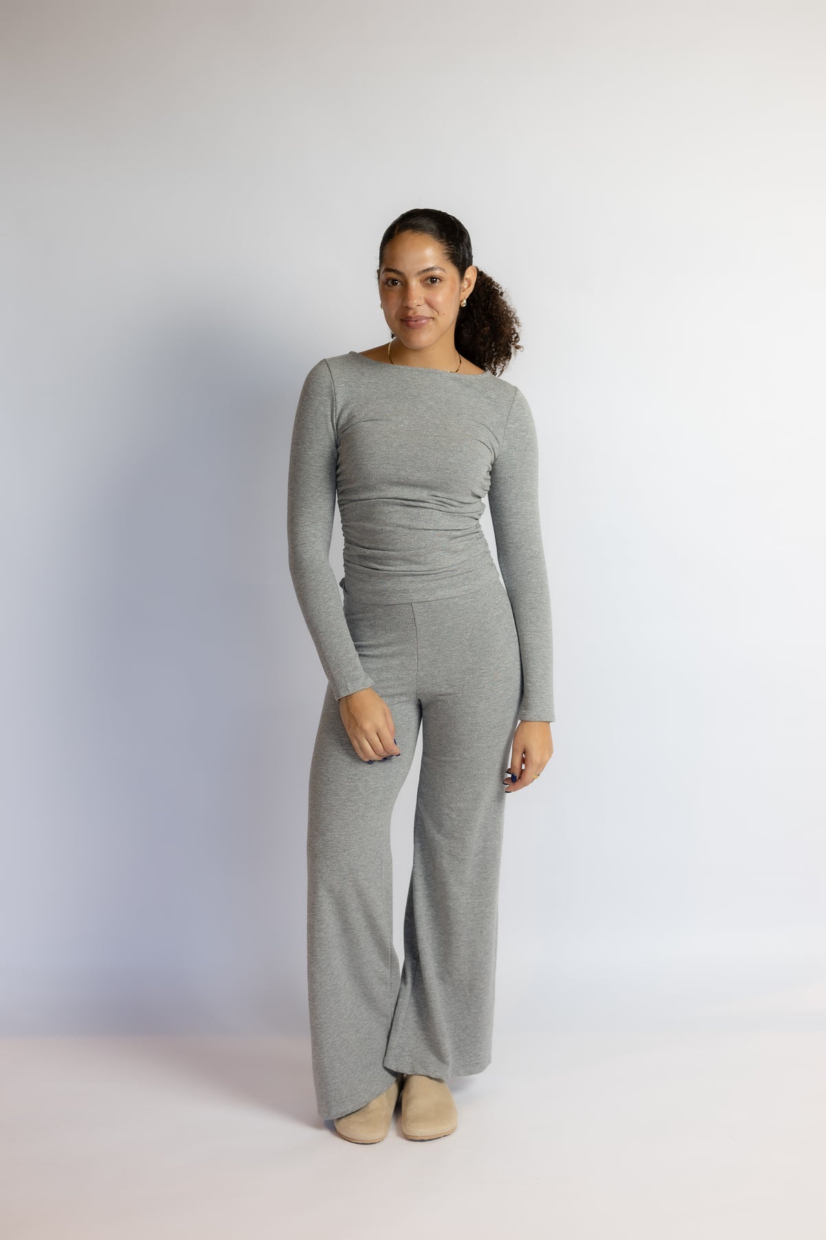 A woman modeling a light gray long-sleeve jumpsuit with a boat neckline and wide-leg pants, standing against a plain light gray studio background.