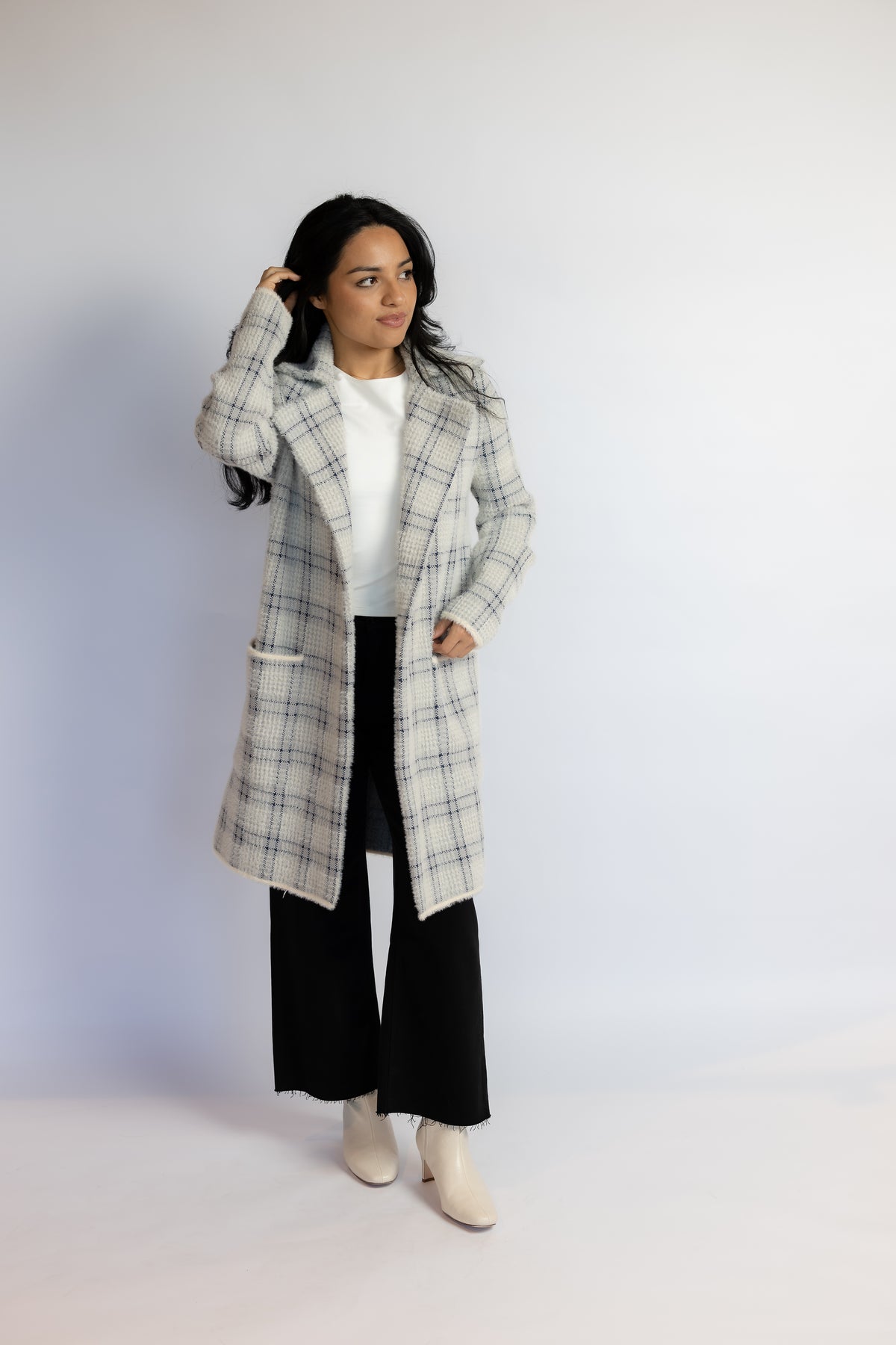 A woman modeling a long white and gray plaid coat over a white top, black wide-leg pants, and off-white ankle boots, standing against a plain light gray studio background.