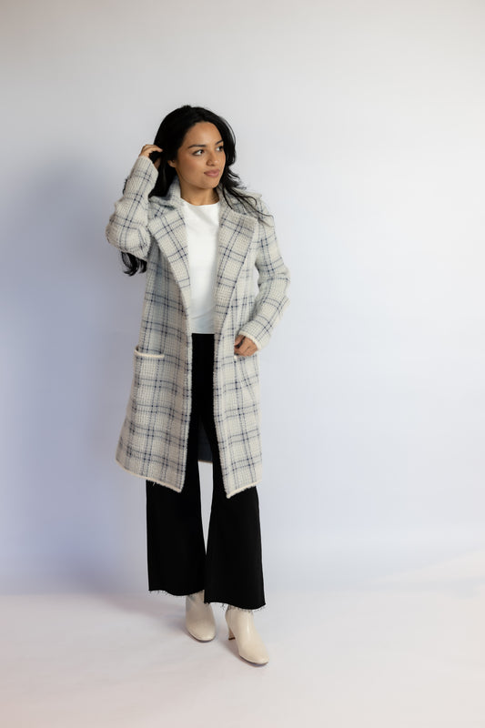 A woman modeling a long white and gray plaid coat over a white top, black wide-leg pants, and off-white ankle boots, standing against a plain light gray studio background.