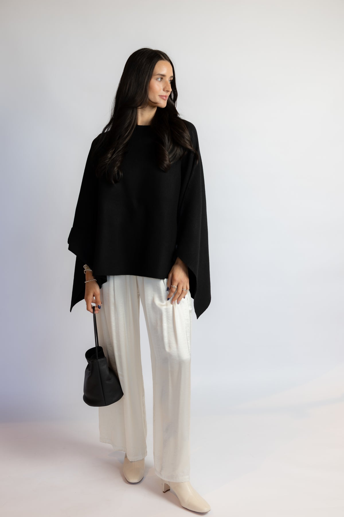 A woman modeling a black draped poncho-style top with wide sleeves, paired with white wide-leg trousers and beige ankle boots, holding a small black handbag against a plain light gray studio background.