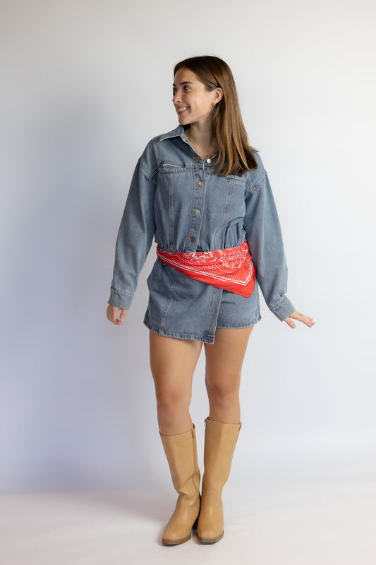A woman modeling a light blue denim shirt-jacket and mini skirt set, styled with a red bandana tied around her waist and tan knee-high boots, against a plain light gray studio background.