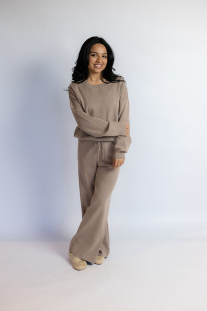 A woman modeling a matching taupe knit lounge set consisting of a long-sleeve sweatshirt and wide-leg pants, standing against a light gray studio background.