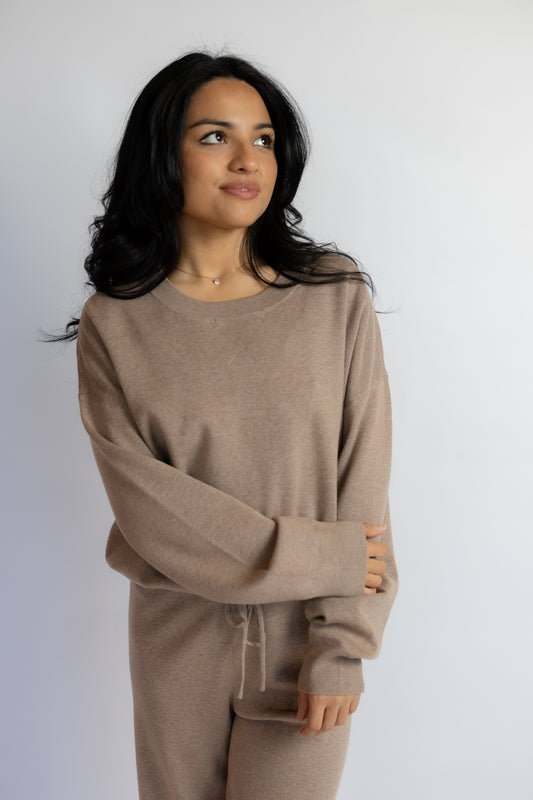 A woman with long dark hair wearing a matching beige knit lounge set, consisting of a long-sleeve top and pants with a drawstring waist, photographed against a plain light gray background.