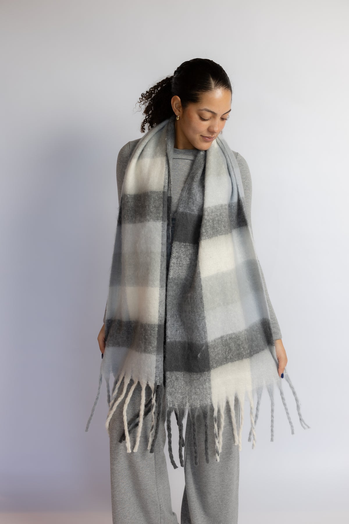 A woman wearing a large gray, white, and black plaid fringed scarf over a gray top and matching gray pants, standing against a light gray studio background.