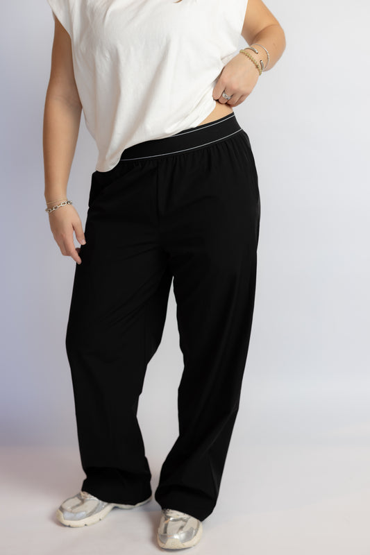 A person wearing black wide-leg pants with a thin white stripe detail along the elastic waistband, paired with a white sleeveless top and white sneakers, shown against a light gray studio background.
