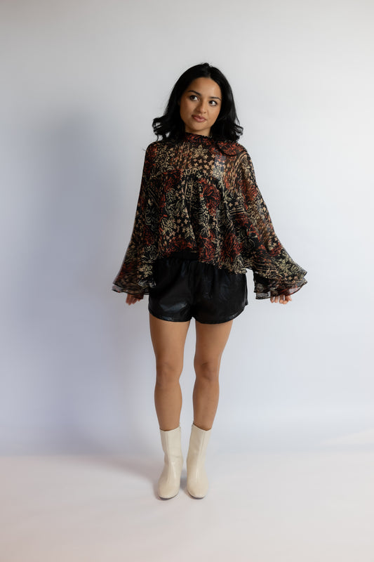 A woman modeling a black sheer long-sleeve blouse with a dense red, gold, and brown floral pattern, paired with black shorts and off-white ankle boots, standing against a plain white studio background.