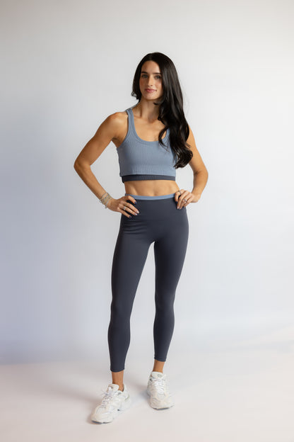 A woman modeling a matching activewear set consisting of a slate blue sports bra and dark gray leggings, paired with white sneakers, against a light gray studio background.