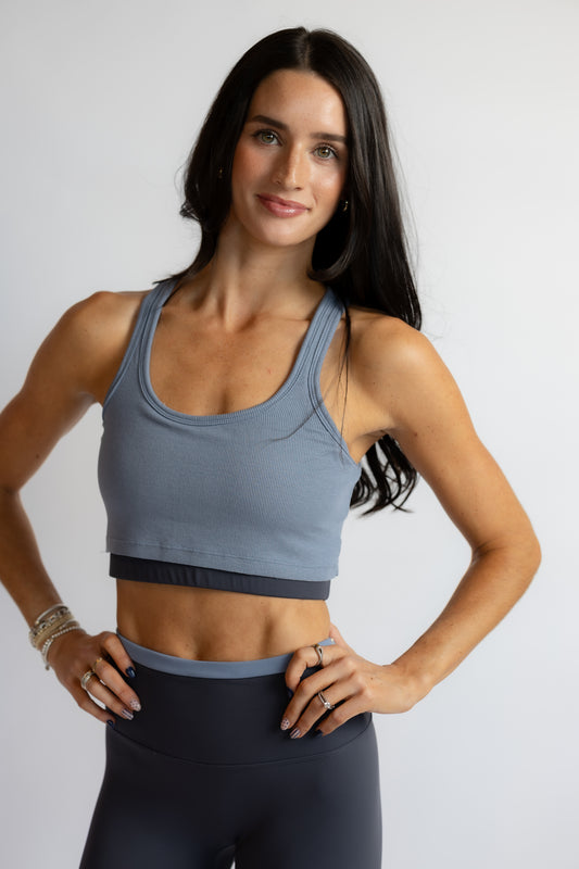 A woman modeling a slate blue ribbed scoop-neck sports bra with a dark navy underband, paired with dark gray leggings, against a light gray studio background.