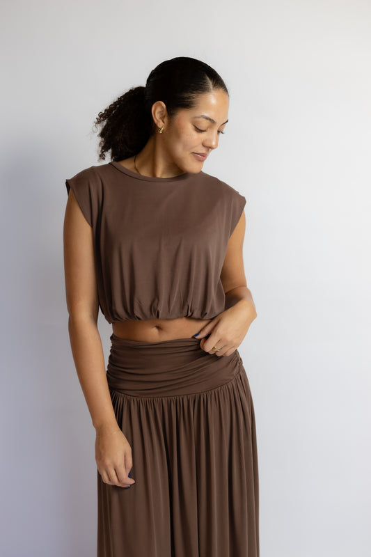 A woman wearing a matching brown two-piece set consisting of a sleeveless cropped top and a high-waisted pleated skirt, standing against a plain light gray background.