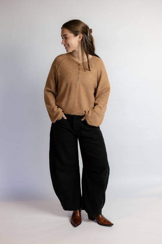 A woman modeling a tan knit henley sweater and black wide-leg pants against a plain light gray studio background.
