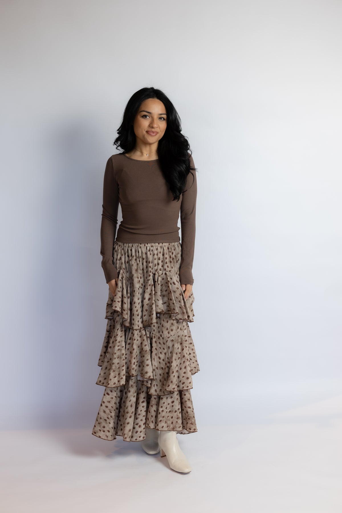 A woman modeling a brown long-sleeve knit top paired with a beige and brown patterned tiered maxi skirt and white ankle boots, standing against a light gray studio backdrop.