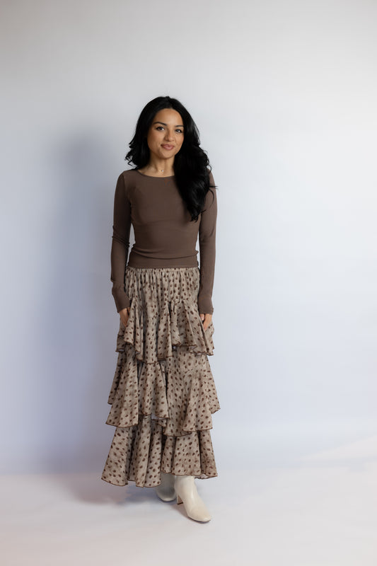 A woman modeling a brown long-sleeve knit top paired with a beige and brown patterned tiered maxi skirt and white ankle boots, standing against a light gray studio backdrop.