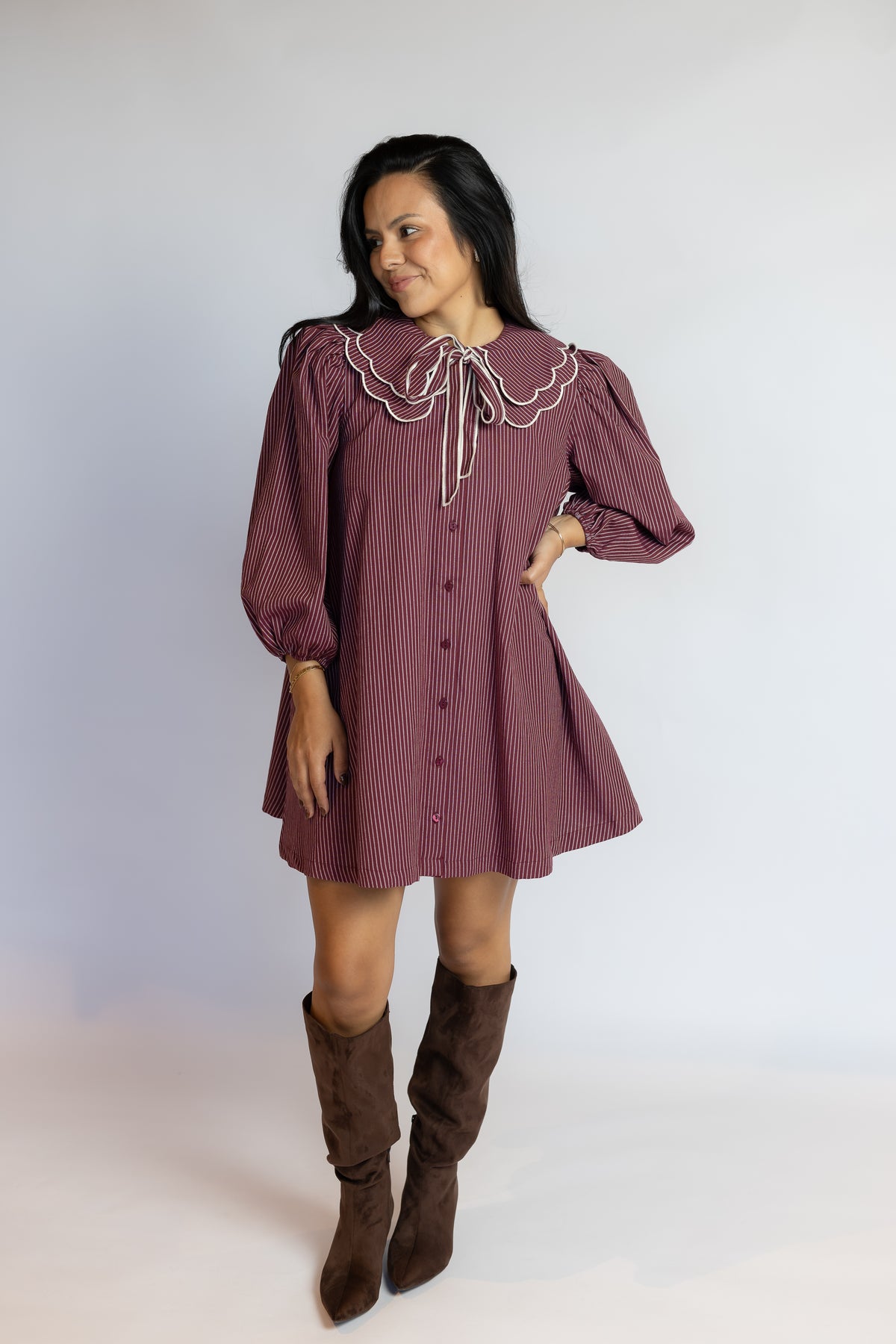 A woman modeling a mauve-colored mini dress with a large ruffled collar, white trim, and brown knee-high boots against a plain light gray studio background.