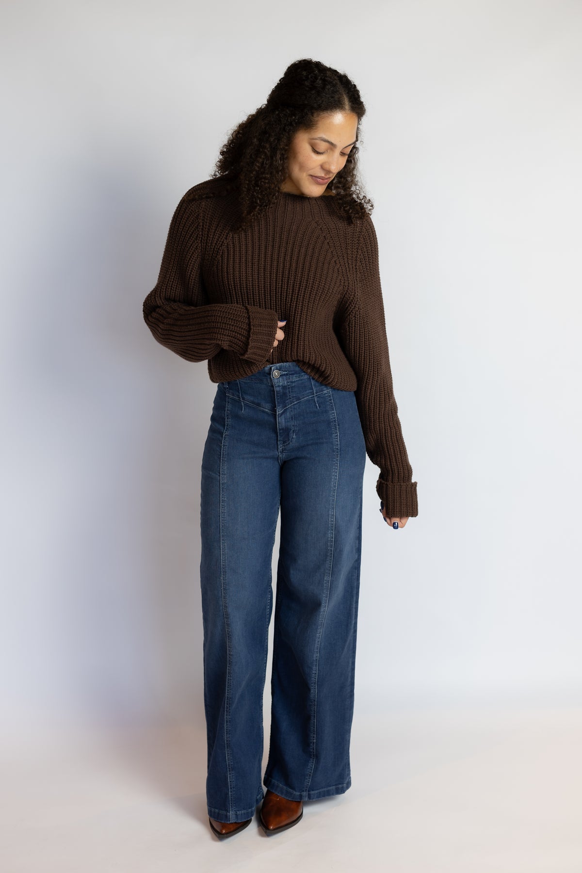 A woman modeling a dark brown ribbed knit sweater and high-waisted, wide-leg blue jeans against a plain white studio background.