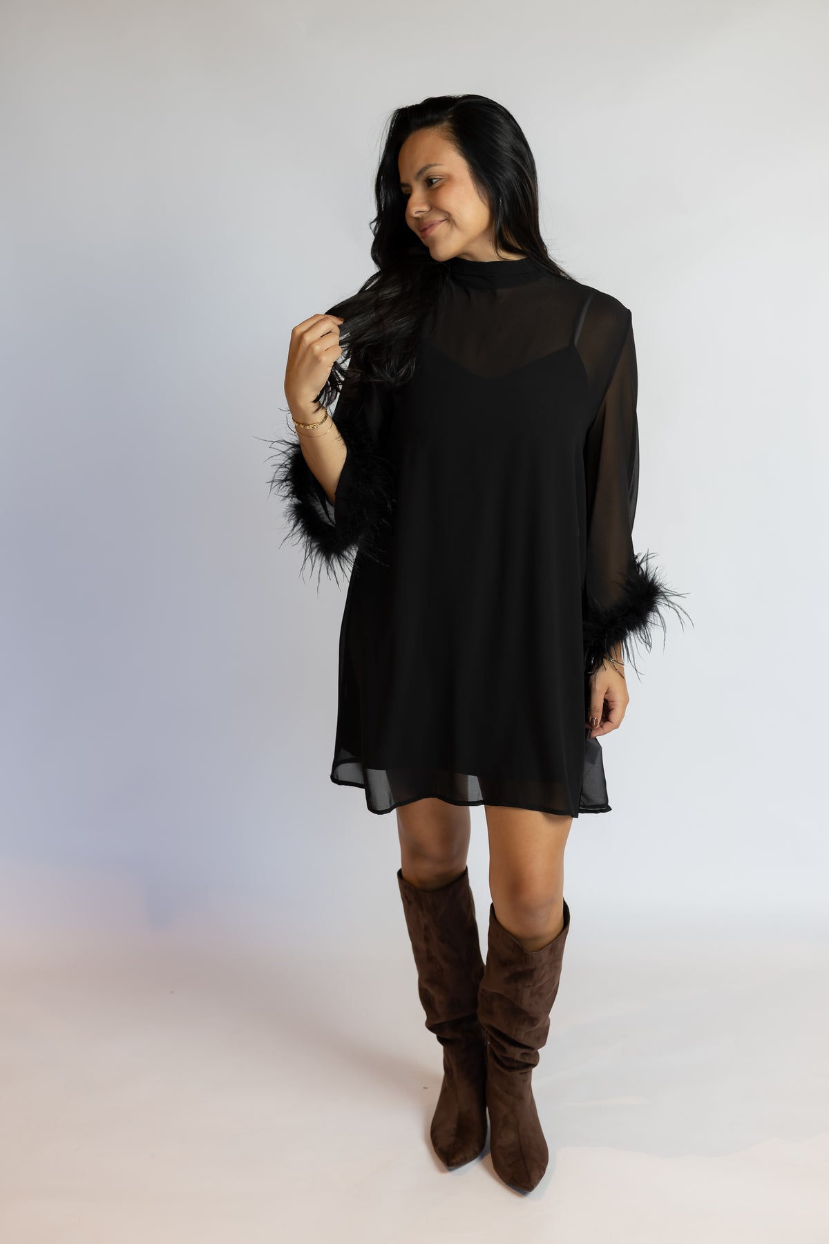 A woman modeling a black sheer long-sleeve mini dress with black feather trim on the sleeves, paired with brown suede knee-high boots, against a light gray studio background.