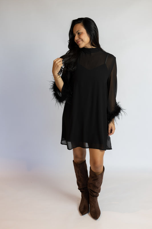 A woman modeling a black sheer long-sleeve mini dress with black feather trim on the sleeves, paired with brown suede knee-high boots, against a light gray studio background.