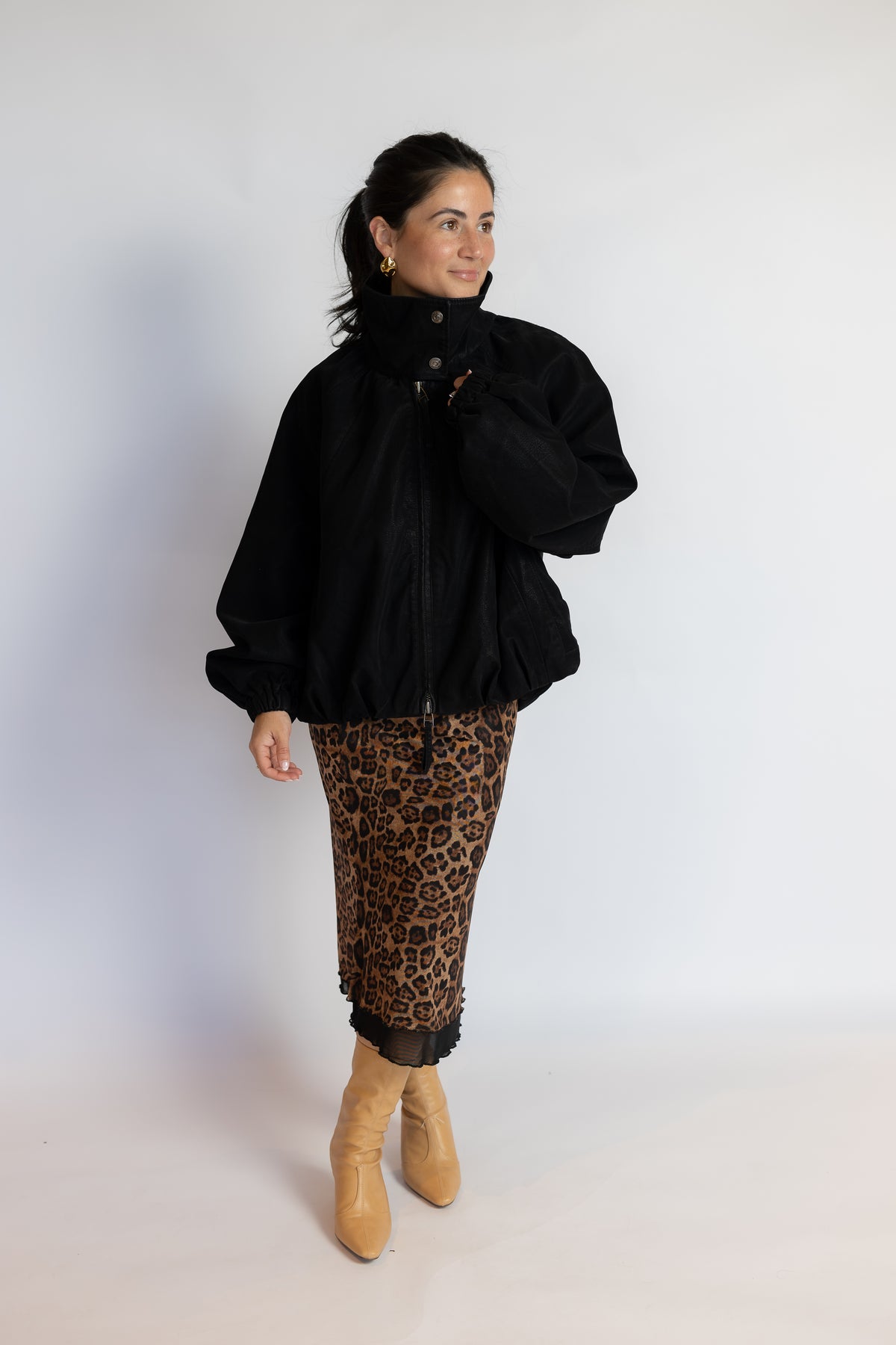 A woman modeling a black oversized puffer jacket with a high collar, paired with brown and black leopard print pants and tan ankle boots, standing against a plain white studio background.