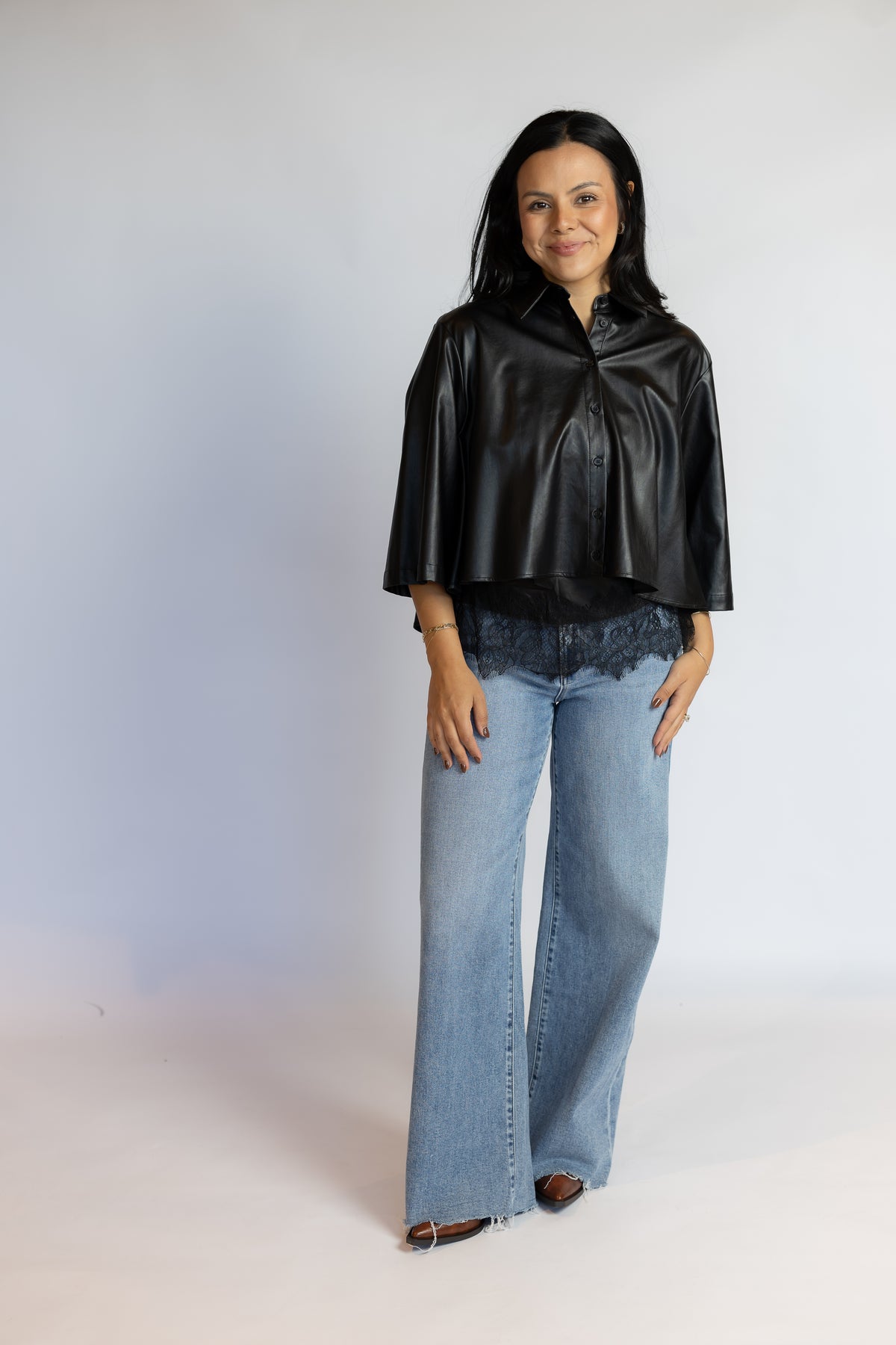 A woman modeling a black faux-leather cropped cape jacket with wide three-quarter sleeves, paired with light-wash wide-leg jeans and brown flat shoes, standing against a light gray studio background.