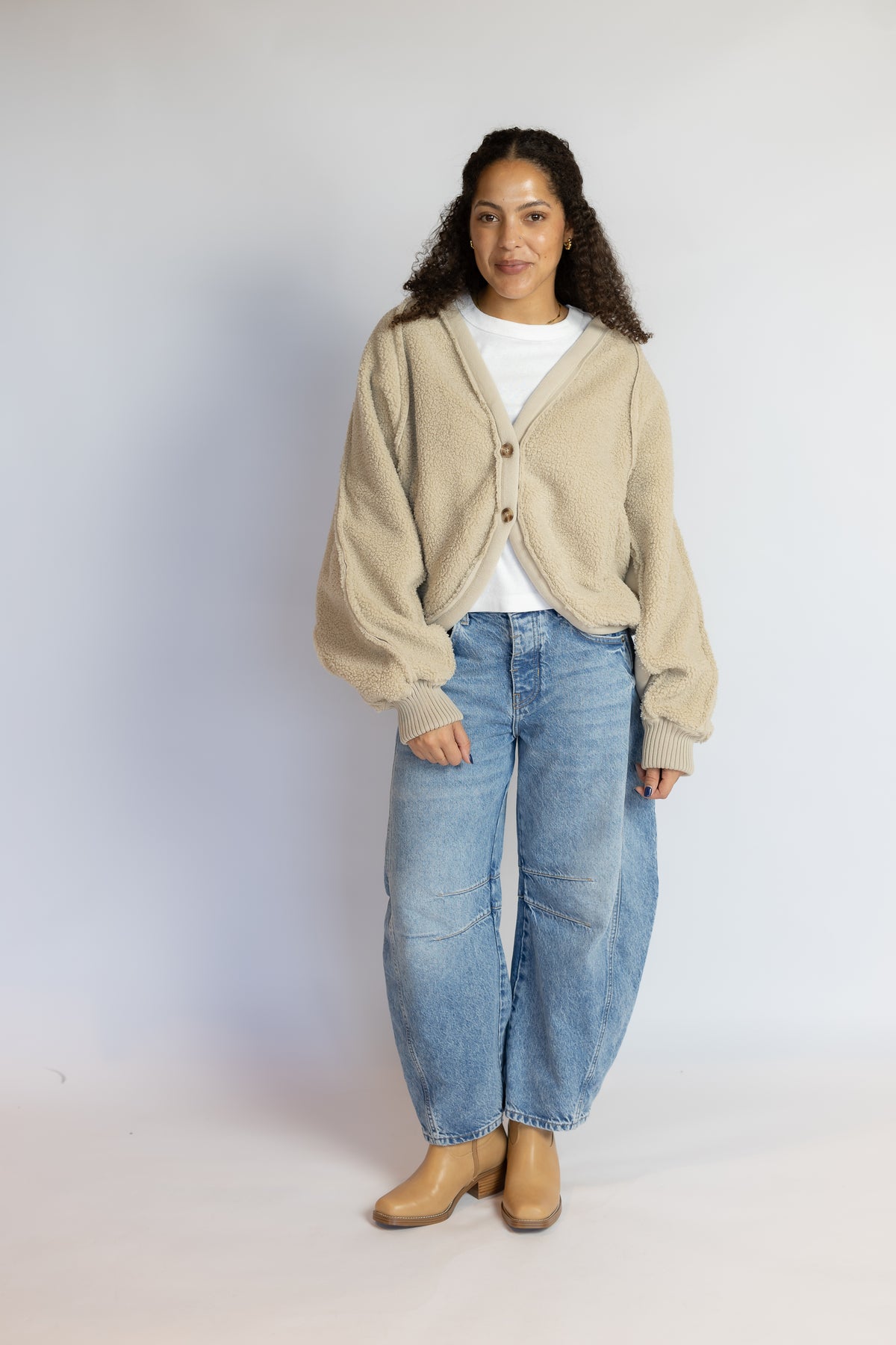 A woman modeling a beige cropped cardigan with balloon sleeves, worn over a white t-shirt and paired with wide-leg blue jeans and tan ankle boots, standing against a light gray studio background.
