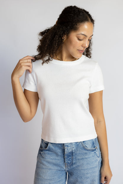 A woman with curly dark hair wearing a white short-sleeve crewneck t-shirt and light blue jeans, standing against a light gray studio background.