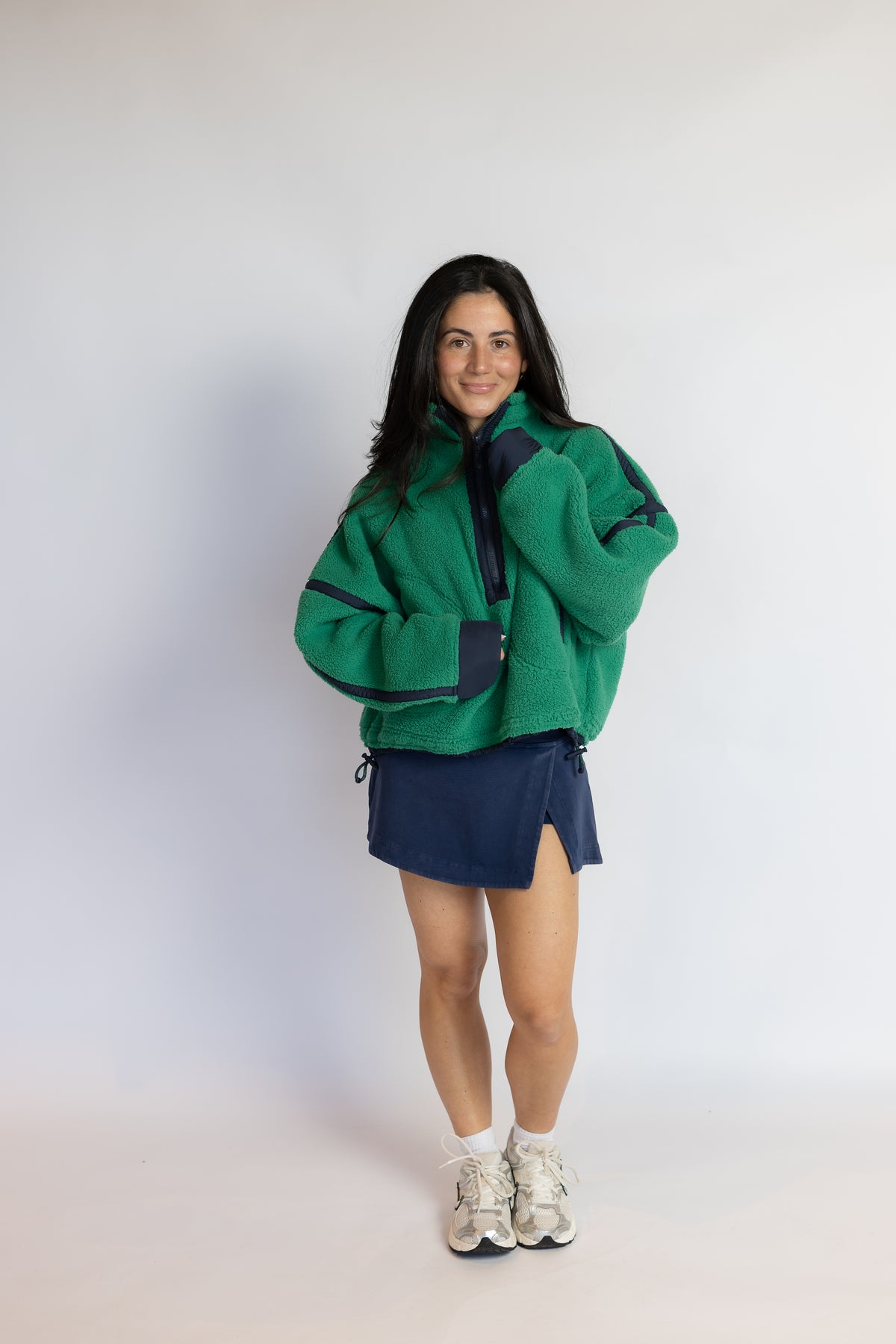 A woman modeling a green fleece zip-up jacket with dark navy panels, paired with a navy mini skirt and white sneakers, standing against a plain light gray studio background.