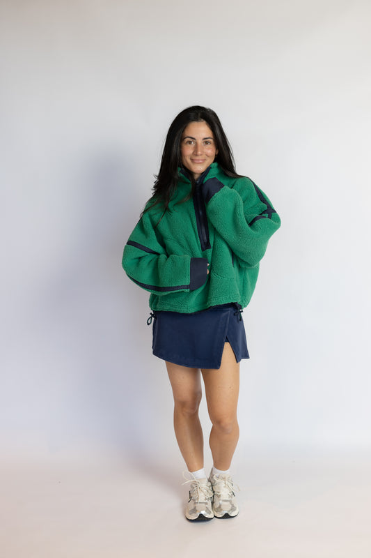 A woman modeling a green fleece zip-up jacket with dark navy panels, paired with a navy mini skirt and white sneakers, standing against a plain light gray studio background.