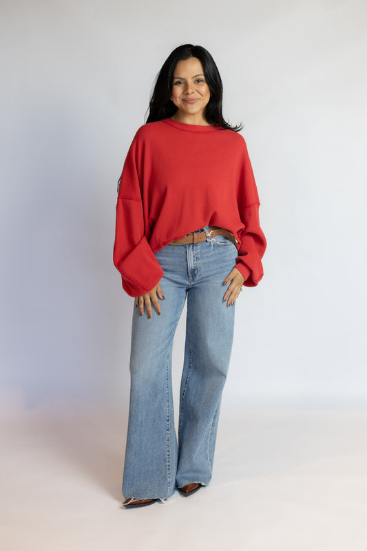 A woman modeling a bright red cropped long-sleeve knit sweater paired with light-wash blue flare jeans, standing against a plain light gray studio background.