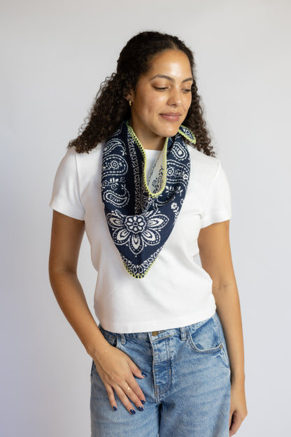A woman with curly dark hair wearing a white t-shirt and blue jeans, modeling a navy blue paisley bandana scarf with lime green trim against a light gray background.