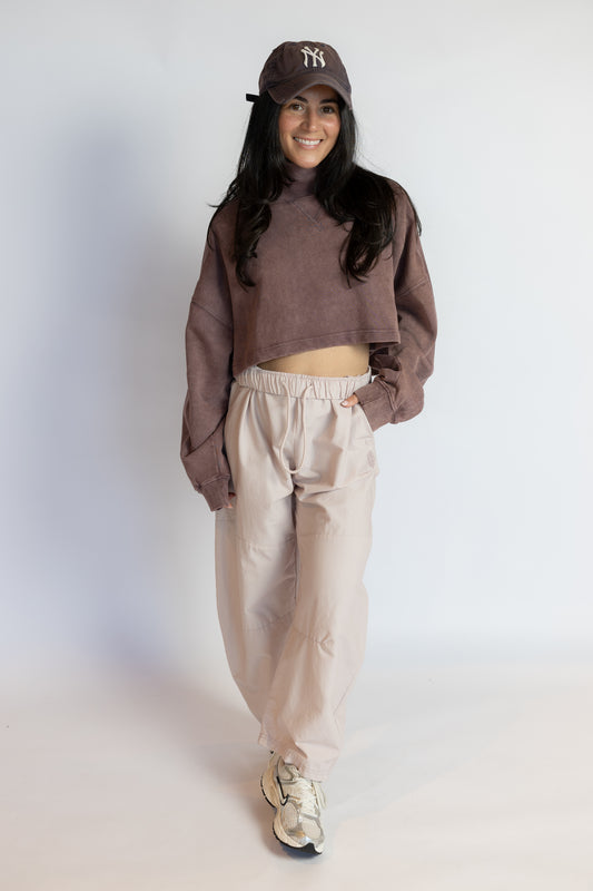 A woman modeling a brown cropped turtleneck sweatshirt, beige baggy sweatpants, a brown New York Yankees baseball cap, and white sneakers against a plain white studio background.