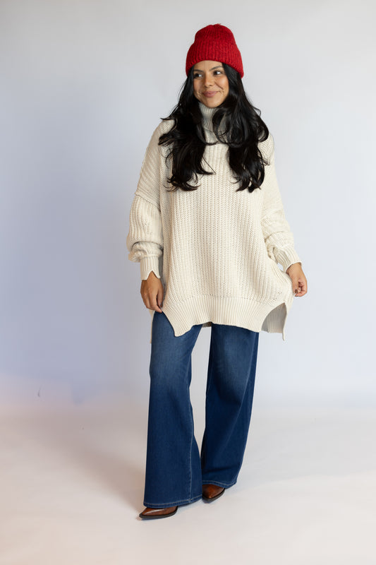 A woman modeling a cream ribbed knit turtleneck sweater, blue wide-leg jeans, a red beanie, and brown shoes against a light gray studio background.