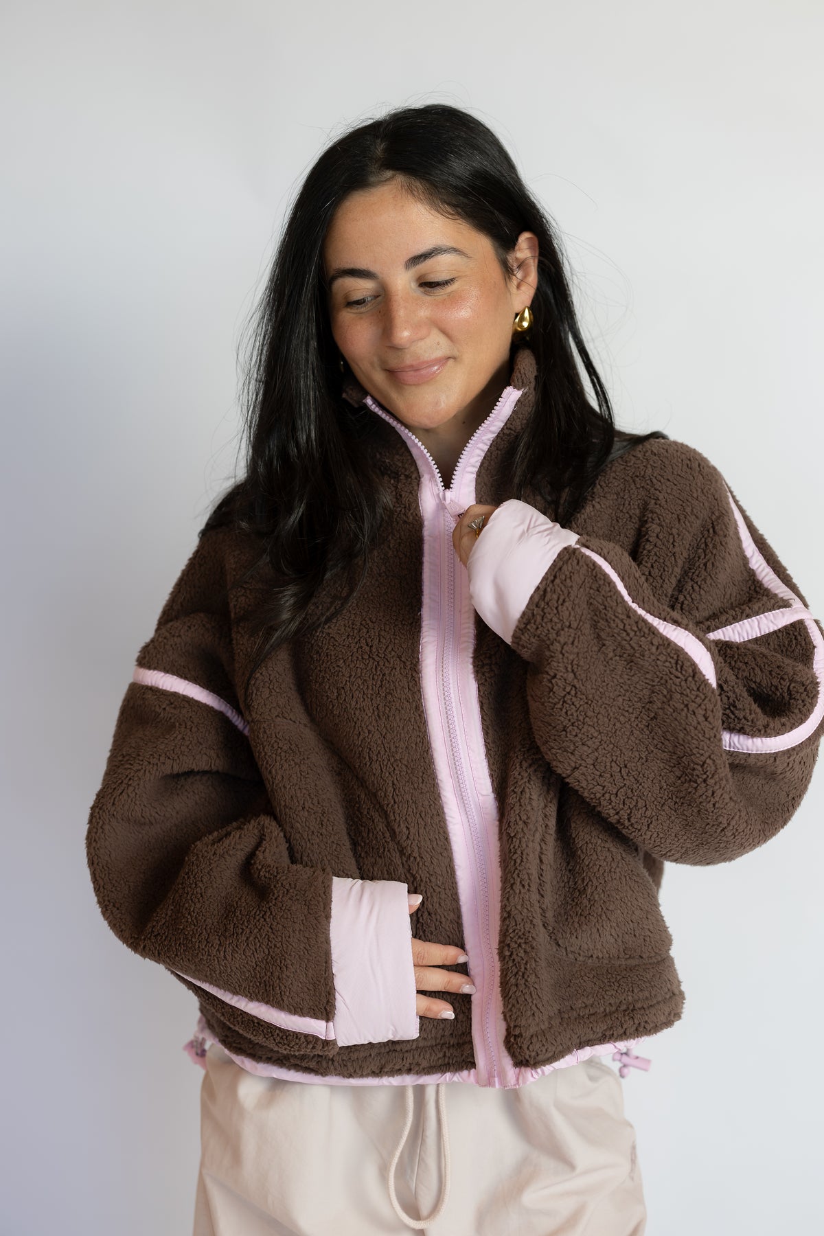 A woman wearing a brown sherpa fleece zip-up jacket with light pink trim and a light pink zipper, paired with cream drawstring pants, set against a plain light gray background.