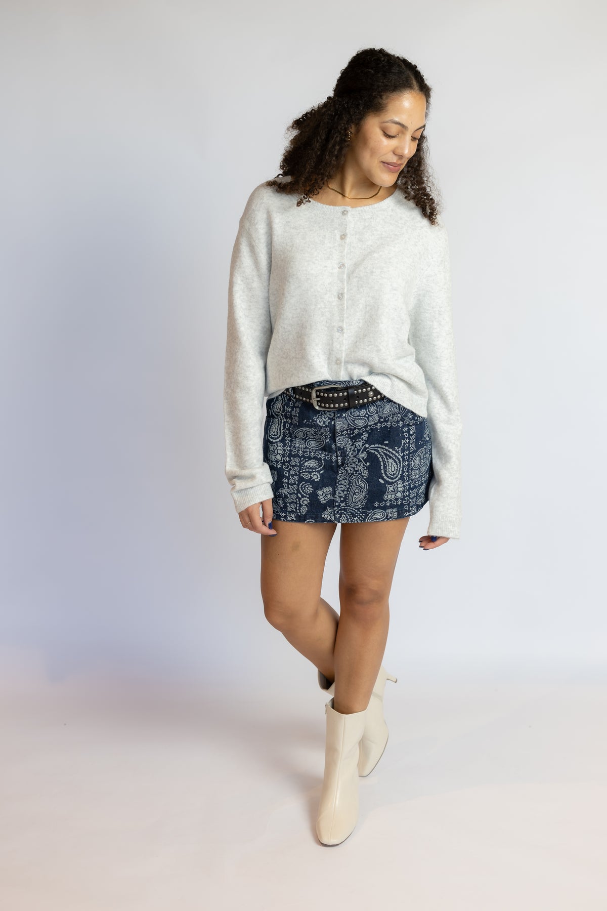 A woman modeling a light gray knit long-sleeve sweater, a blue patterned denim mini skirt, and off-white ankle boots, standing against a light gray studio background.