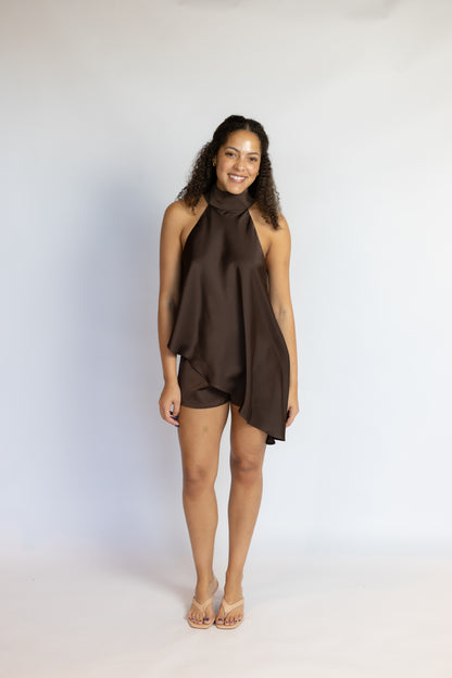A woman modeling a sleeveless, high-neck, asymmetrical mini dress in a dark brown satin-like fabric, standing against a plain light gray studio background.