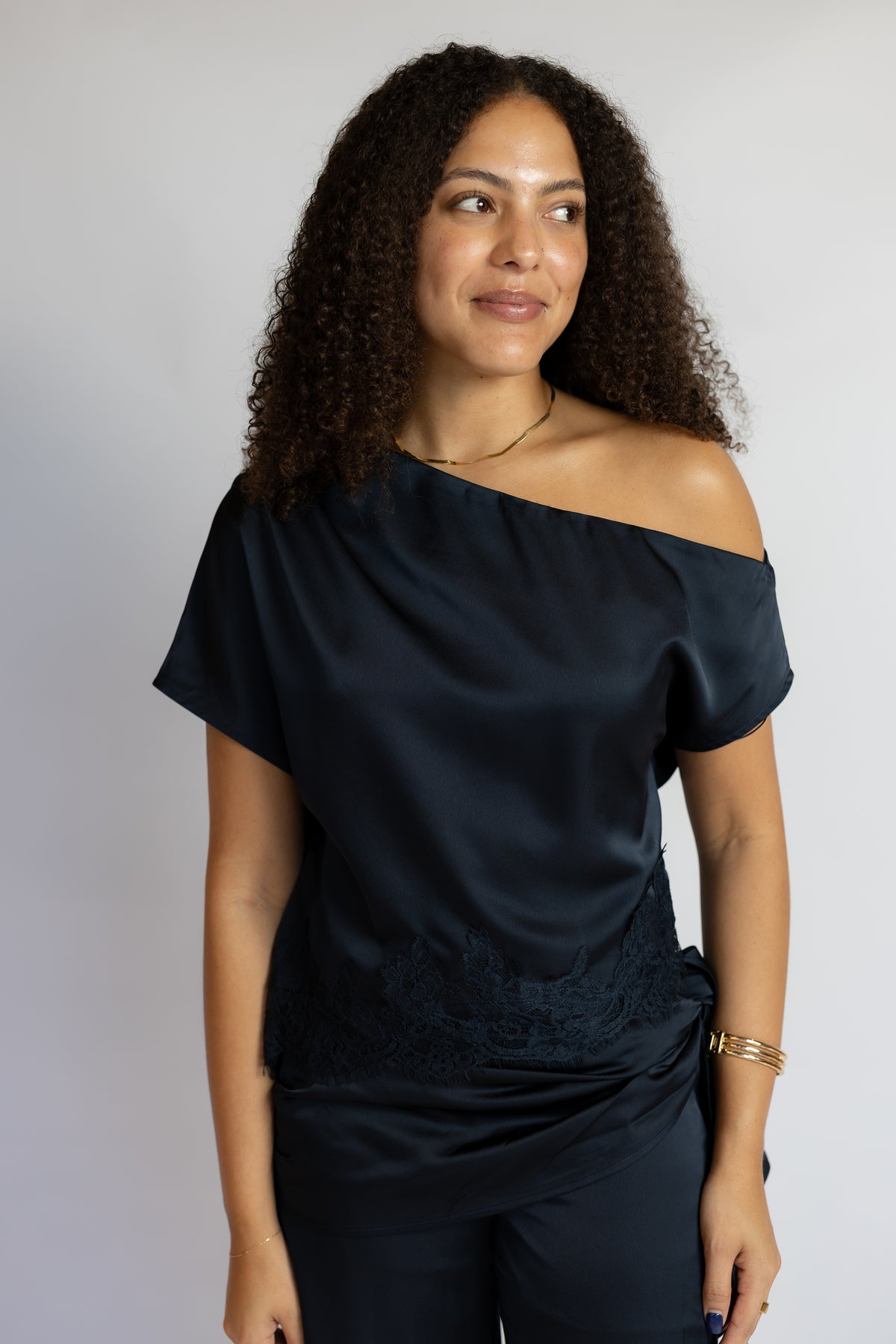 A woman with curly dark hair wearing a dark navy off-the-shoulder satin top with lace detailing at the waist, paired with matching dark pants, against a plain light gray background.
