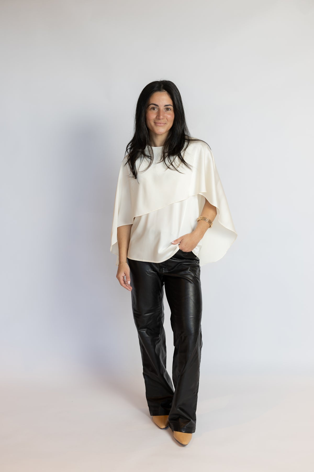 A woman modeling a white asymmetrical draped top and black leather pants against a light gray studio background.