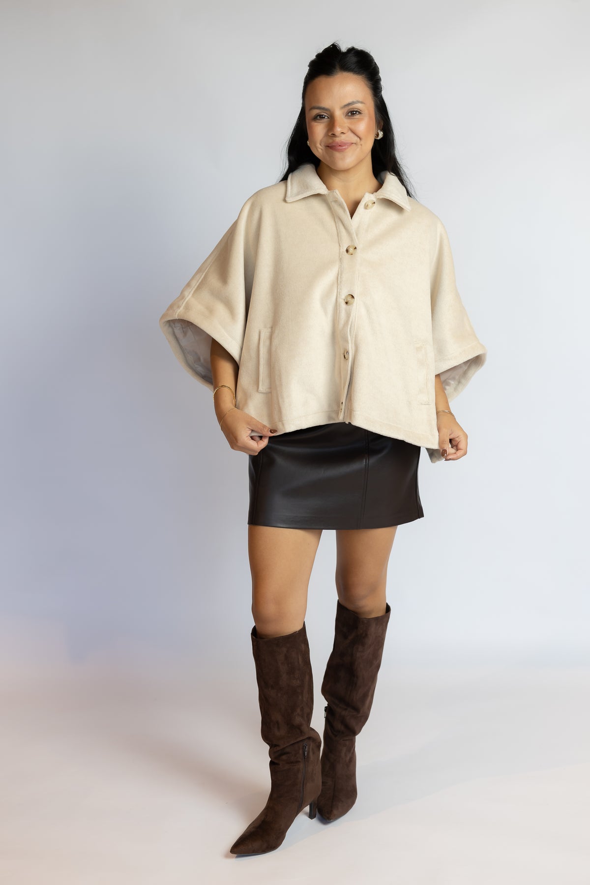 A woman modeling a cream-colored short-sleeved button-up shirt jacket with a collar, paired with a black mini skirt and brown knee-high boots, against a light gray studio background.