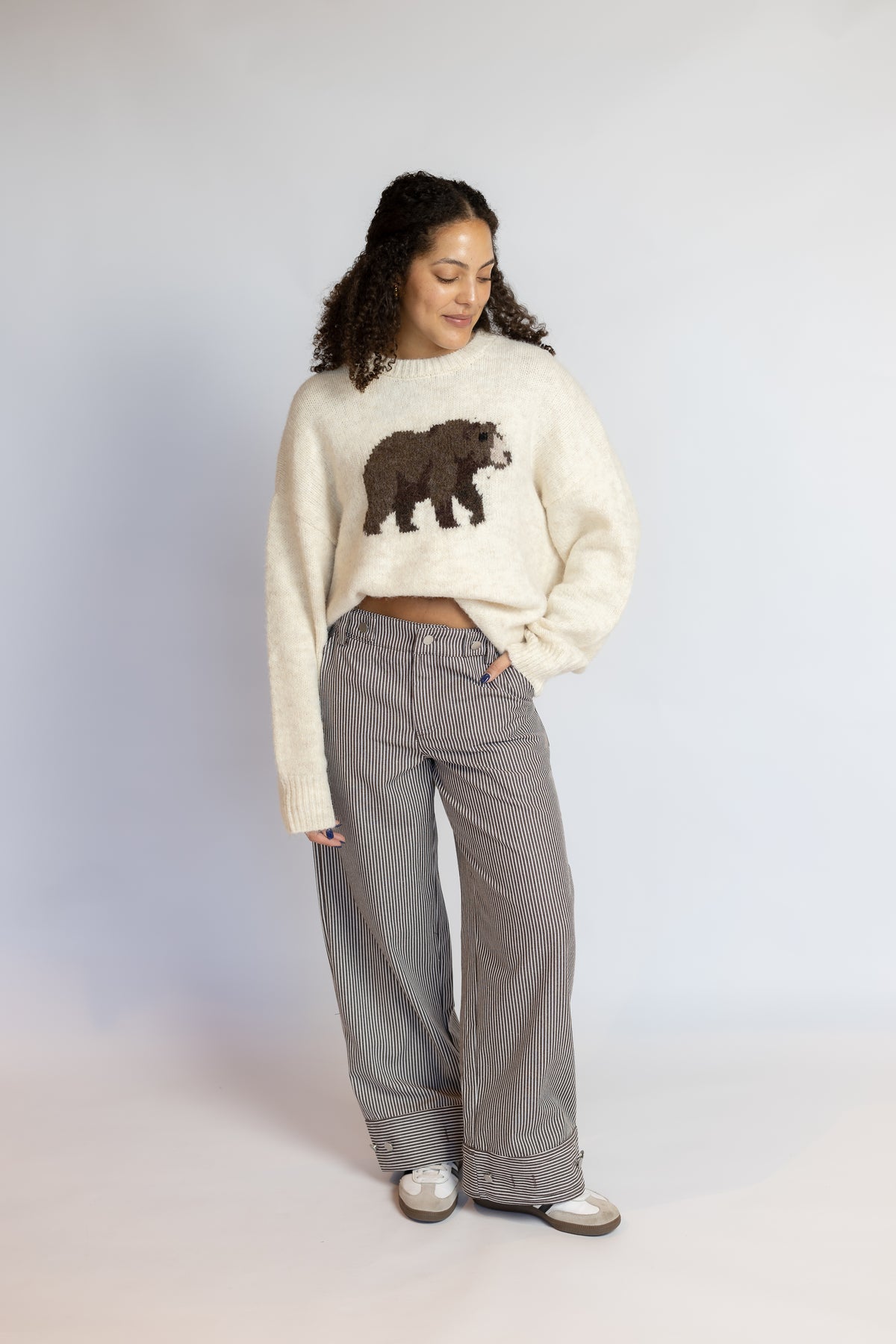 A woman stands against a light gray studio background wearing a cream knit sweater with a brown bear graphic on the chest, gray wide-leg pants, and white sneakers.