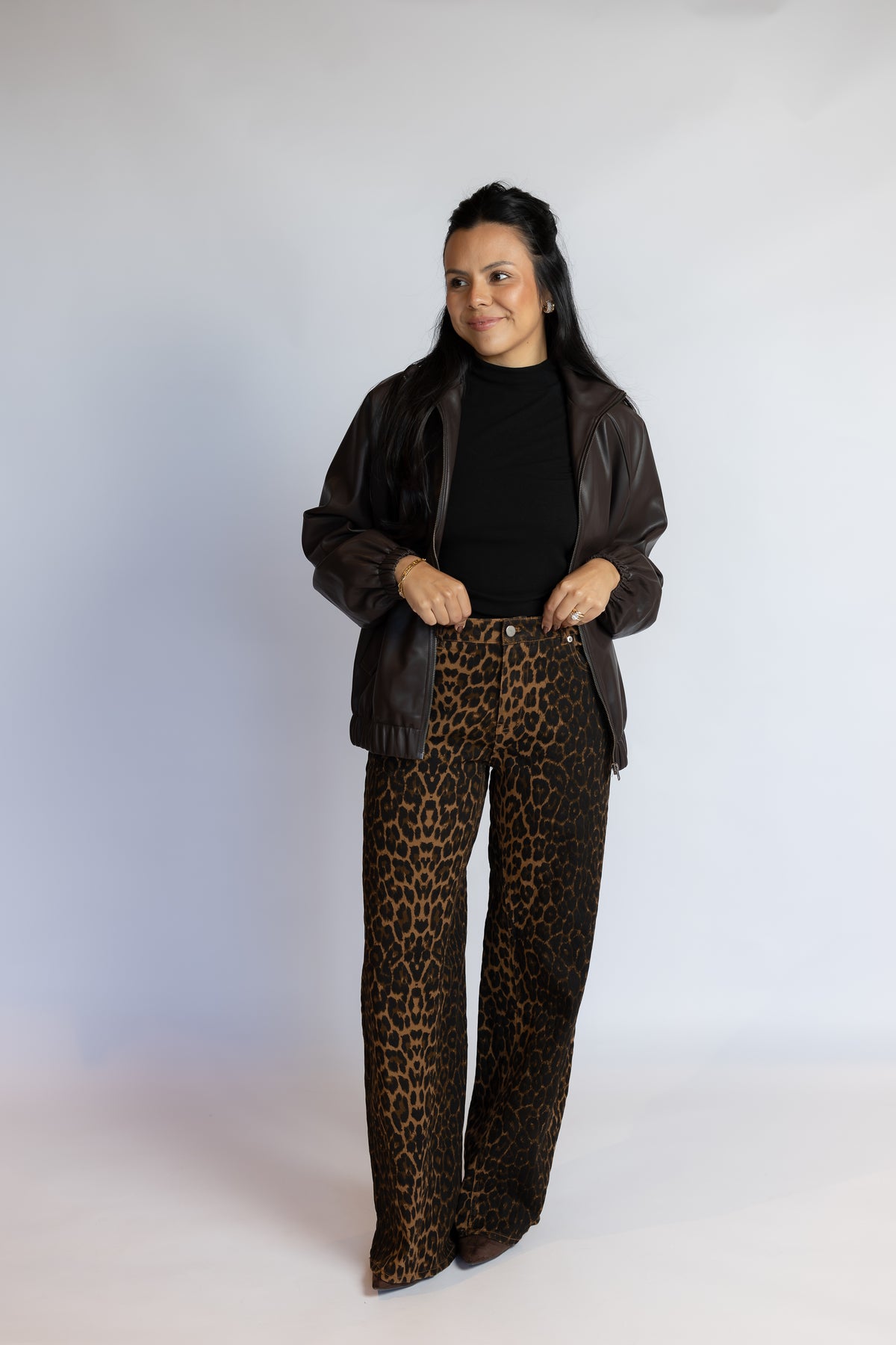A woman modeling a dark brown leather jacket, black turtleneck, and brown leopard-print wide-leg pants against a plain light gray studio background.