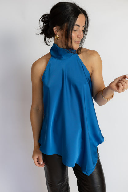 A woman wearing a bright blue satin halter top and black leather pants, standing against a plain light gray studio background.
