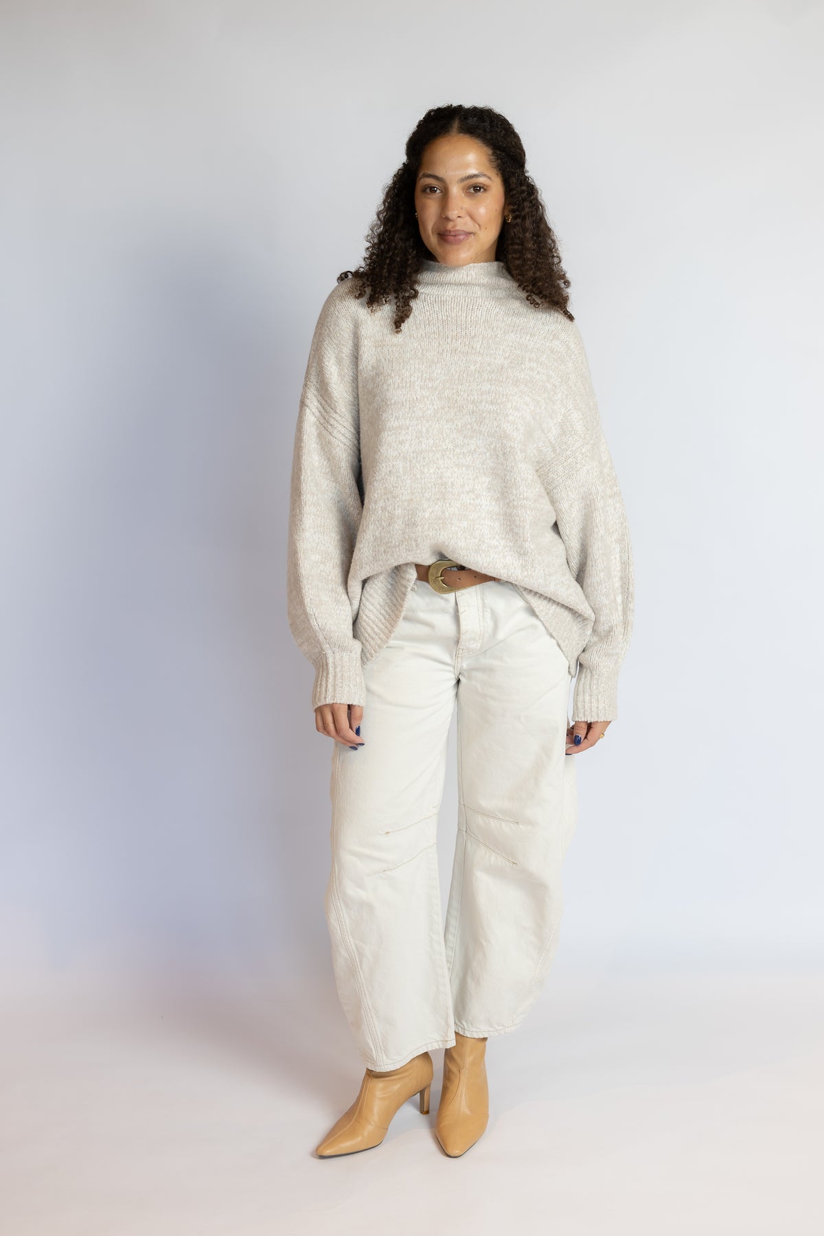 A woman modeling a light beige knit mock-neck sweater, paired with off-white wide-leg jeans and tan ankle boots, standing against a light gray studio background.