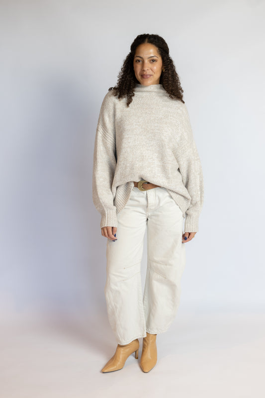 A woman modeling a light beige knit mock-neck sweater, paired with off-white wide-leg jeans and tan ankle boots, standing against a light gray studio background.