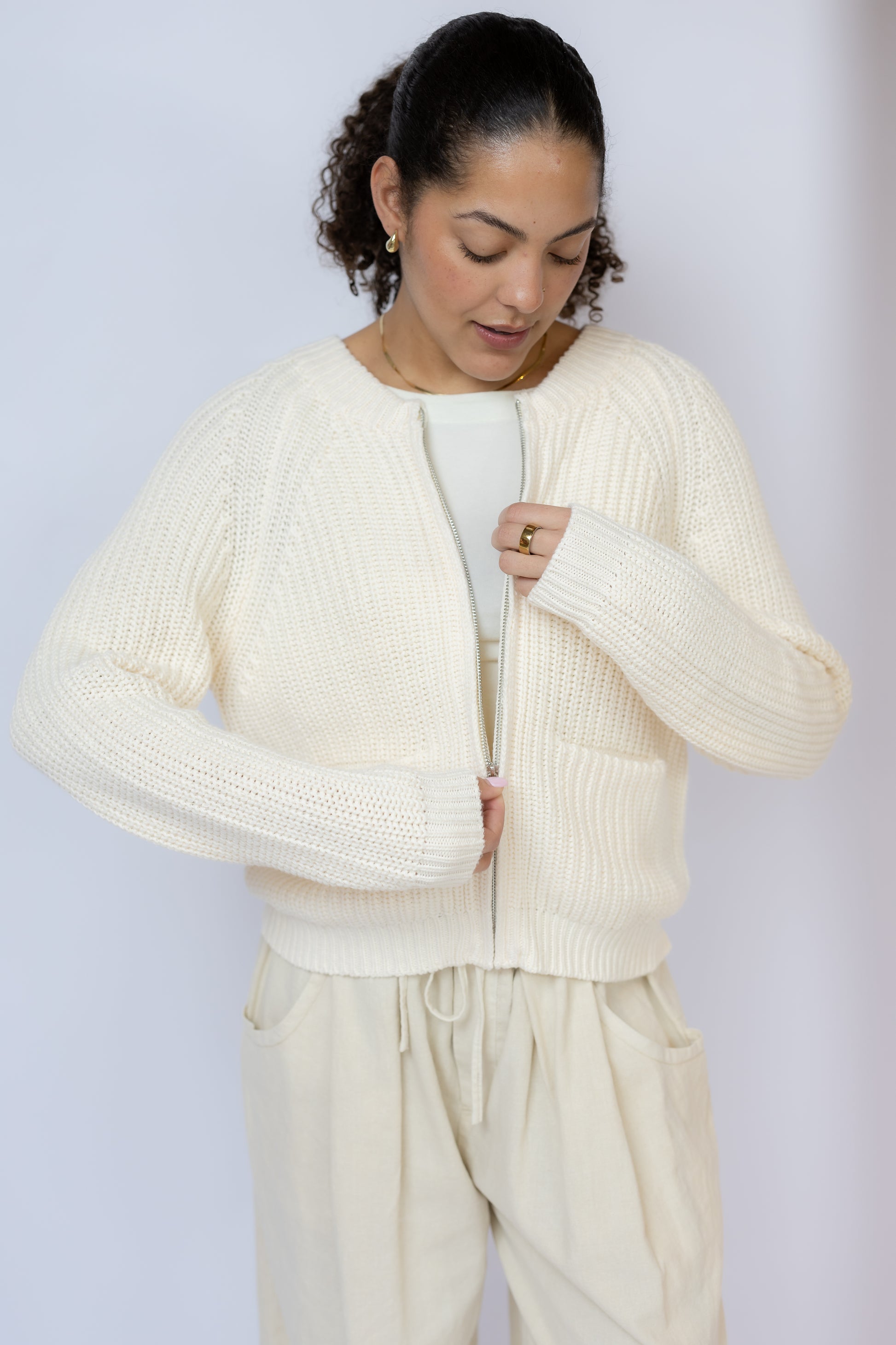 Woman wearing a cream-colored cardigan over a white top and pants against a light gray background