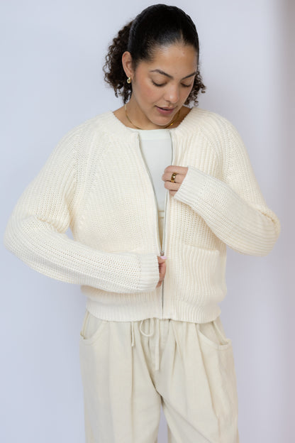 Woman wearing a cream-colored cardigan over a white top and pants against a light gray background