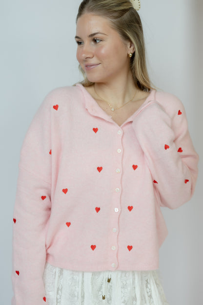 Woman wearing a pink cardigan with red heart patterns against a plain background