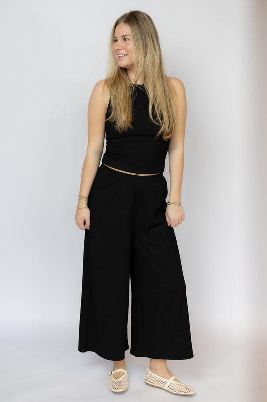 Woman wearing a black sleeveless top and wide-leg pants against a white background